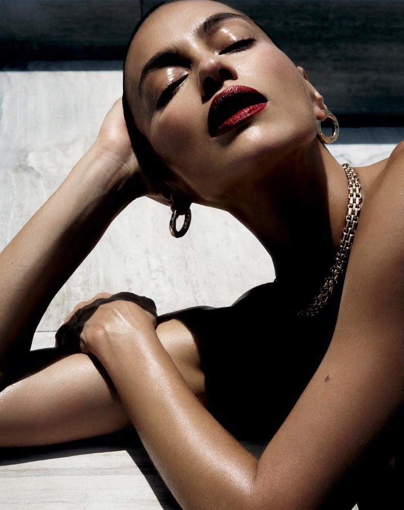 Model Irina Shayk wearing gold hoop earrings and a chain necklace with dark red lipstick under bright sunlight.