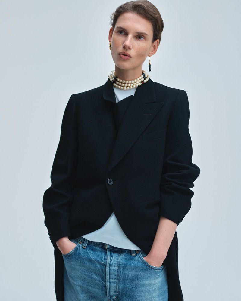 Model Giedre Dukauskaite wearing a black tailored blazer, pearl choker, and blue denim jeans against a pale studio background.