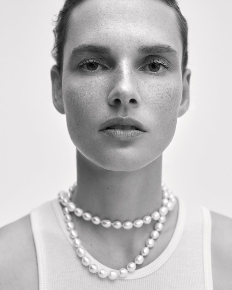 Close-up portrait of model Giedre Dukauskaite wearing a layered pearl necklace and white tank top against a plain studio background.