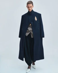 Model Giedre Dukauskaite standing in a full-length navy coat with a gold brooch and white heels against a pale studio backdrop.