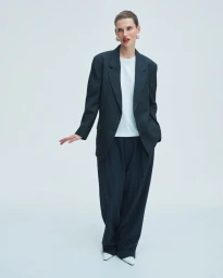 Model Giedre Dukauskaite wearing an oversized dark suit, white top, and pointed shoes against a light grey seamless studio backdrop.
