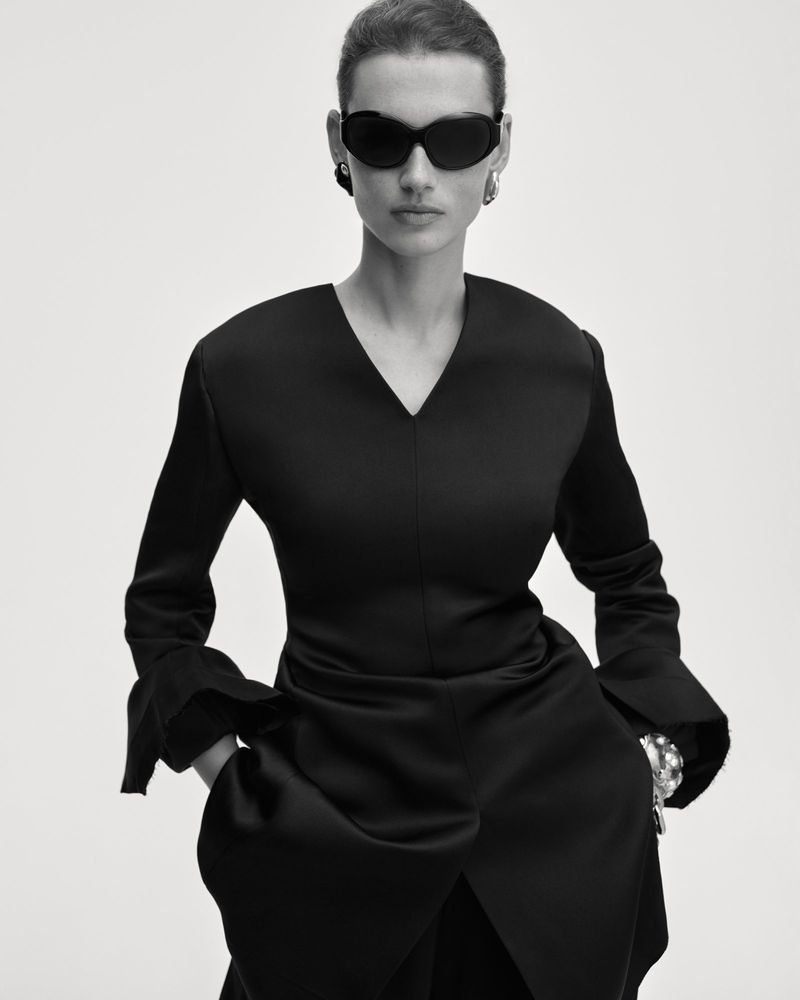 Model Giedre Dukauskaite posing in dark sunglasses and a black v-neck long-sleeve garment against a white studio backdrop.