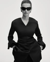 Model Giedre Dukauskaite posing in dark sunglasses and a black v-neck long-sleeve garment against a white studio backdrop.