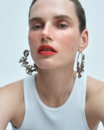 Close-up beauty portrait of model Giedre Dukauskaite wearing bold red lipstick, large floral hoop earrings, and a white tank top.