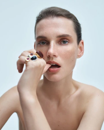 Close-up portrait of model Giedre Dukauskaite with bare shoulders holding statement rings near her lips against a pale background