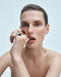 Close-up portrait of model Giedre Dukauskaite with bare shoulders holding statement rings near her lips against a pale background