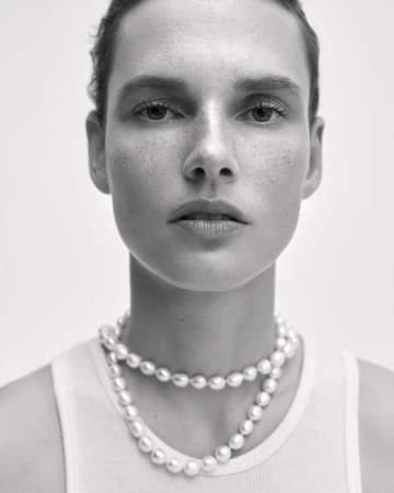Close-up portrait of model Giedre Dukauskaite wearing a layered pearl necklace and white tank top against a plain studio background.