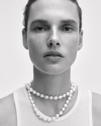 Close-up portrait of model Giedre Dukauskaite wearing a layered pearl necklace and white tank top against a plain studio background.