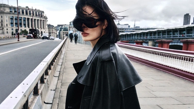 Model wearing a black leather trench coat and sunglasses on an urban bridge overlooking city buildings under an overcast sky.