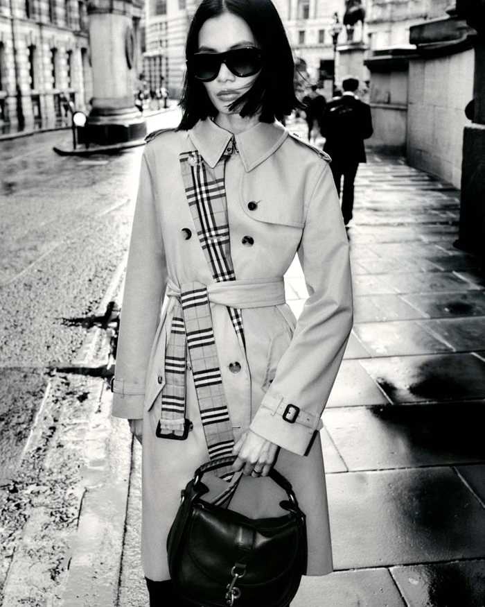 Model in sunglasses wearing a trench coat with plaid scarf and carrying a dark handbag on a wet city street