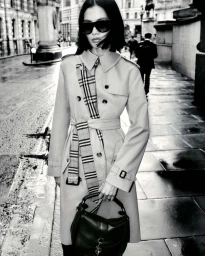Model in sunglasses wearing a trench coat with plaid scarf and carrying a dark handbag on a wet city street