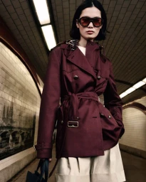 Model wearing dark sunglasses and a burgundy trench coat standing in a tiled subway station