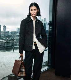Model in black quilted jacket and dark trousers holding a brown handbag against a city skyline view.