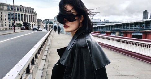 Model in dark sunglasses and black leather coat looking back while standing on a city bridge with traffic and buildings