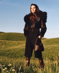 Model standing outdoors in a grassy meadow under a clear blue sky, wearing a furry jacket and carrying a monogram shoulder bag.