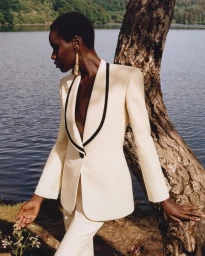 Model wearing a cream suit with black lapel trim standing by a tree trunk near a lake for the Louis Vuitton Fall-Winter 2026 campaign