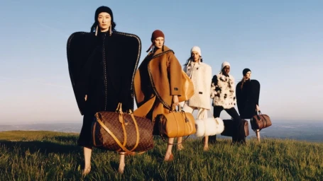Five models in winter apparel holding monogram duffel bags standing in a grassy field under a clear blue sky.