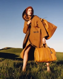 Model in brown hooded coat carrying large tan leather handbags standing in a grassy meadow under a clear blue sky
