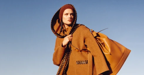 Model in brown hooded coat and beanie carrying a large tan leather shoulder bag against a clear blue sky background