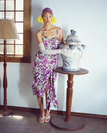 East Asian woman in a draped purple floral dress and white lace gloves posing beside an ornate ceramic urn on a wooden pedestal.