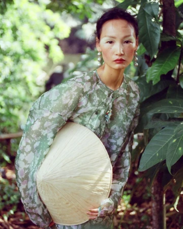 An East Asian woman in a printed green blouse holding a conical straw hat amidst lush tropical greenery.