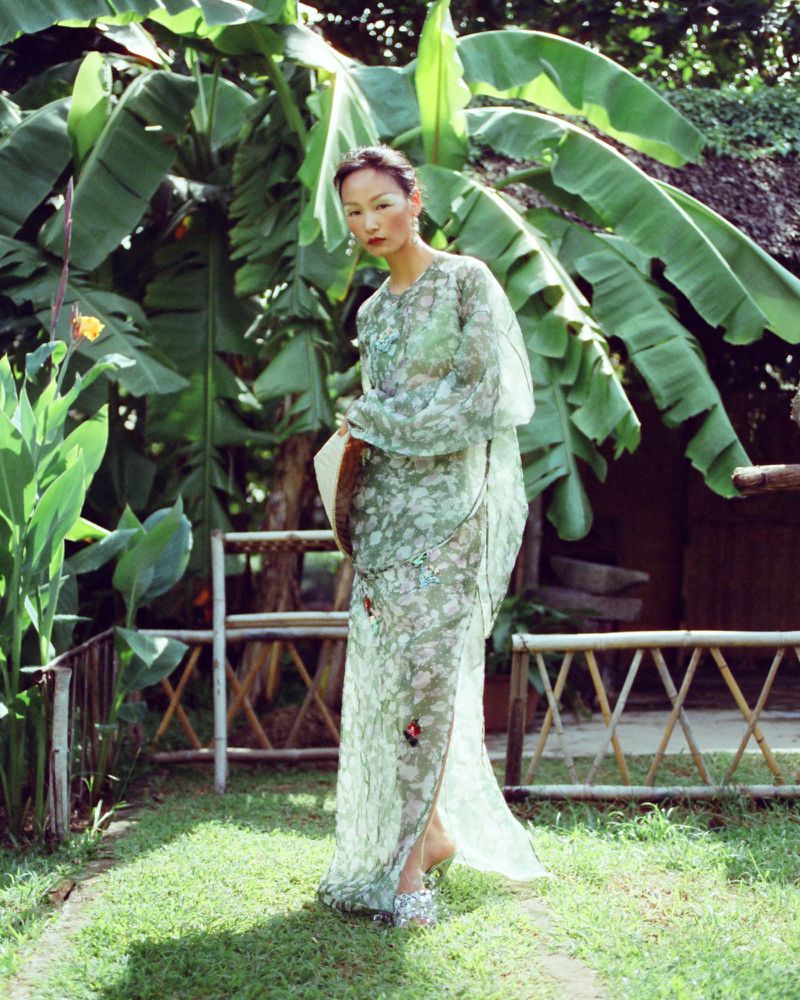 Model wearing a sheer printed sage green gown standing outdoors beside banana leaves and a bamboo fence.