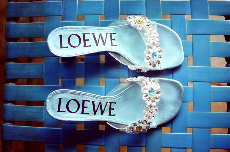 Pair of light blue Loewe slide sandals with crystal floral straps resting on a blue woven lattice surface.