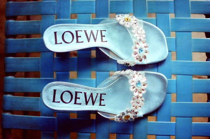 Pair of light blue Loewe slide sandals with crystal floral straps resting on a blue woven lattice surface.