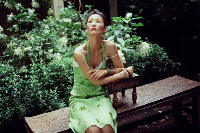 East Asian woman in a light green dress seated on a rustic wooden bench surrounded by lush green foliage and blooming white flowers.