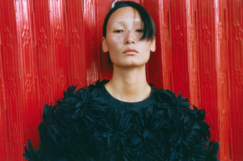 Model with facial freckles wearing a black textured floral appliqué top against a vivid red corrugated shutter.