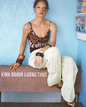 An East Asian woman in a patterned camisole and wide-leg trousers sits on a stone bench against a blue textured wall.