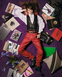Model lying on purple carpet surrounded by magazines, books, and Versace accessories wearing leather vest, white shirt, and red trousers.