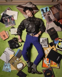 Model lying on a green carpet surrounded by scattered books, magazines, and pillows in a Versace campaign shot.