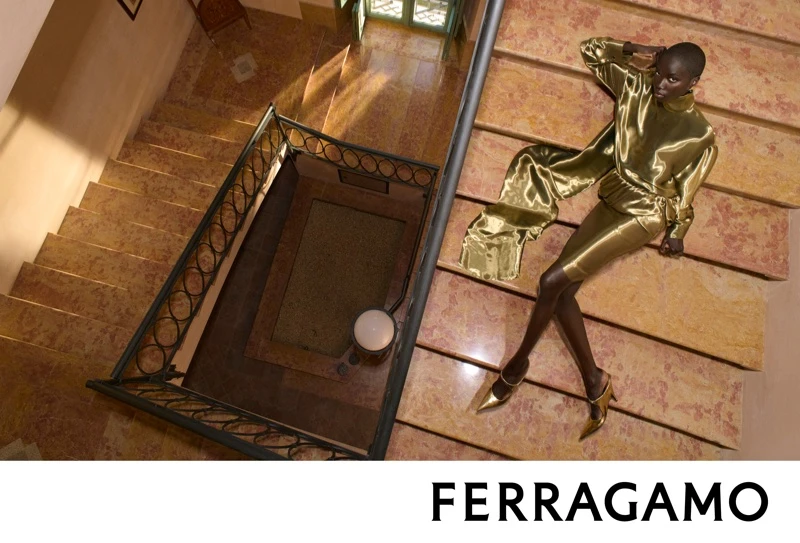 Anok Yai reclining on a grand marble staircase wearing a glossy gold outfit for Salvatore Ferragamo Fall-Winter 2026 campaign