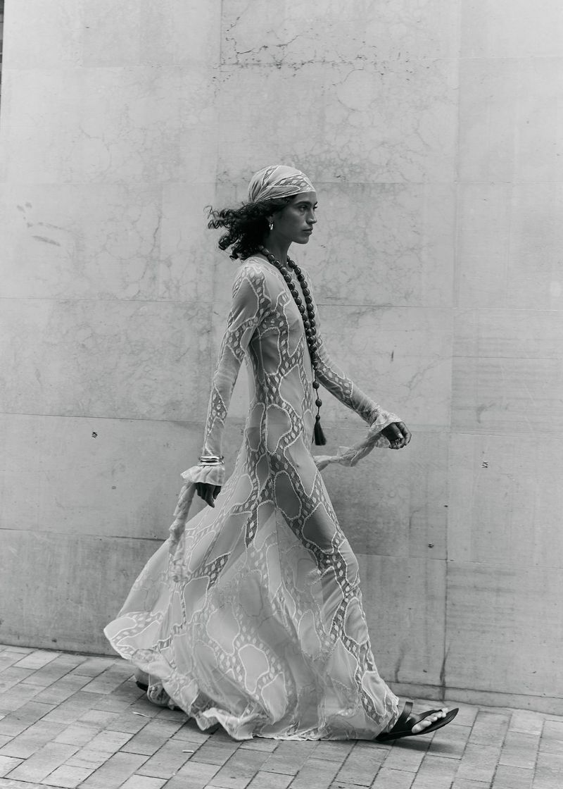 Monochrome fashion photograph of a person walking beside a wall in a sheer patterned maxi dress, headscarf, and sandals.