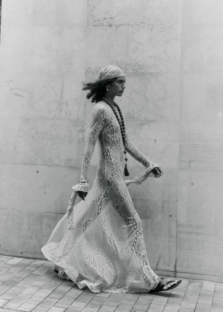 Monochrome fashion photograph of a person walking beside a wall in a sheer patterned maxi dress, headscarf, and sandals.