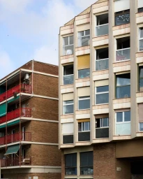Low angle view of multi-story residential apartment buildings with faceted windows, roller blinds, and red balconies under a light blue sky.