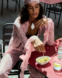Woman in sheer pink patterned outfit and matching headscarf seated at an outdoor cafe table with a bowl of olives.