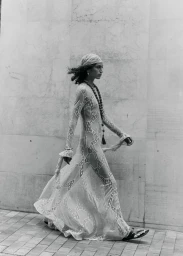 Black and white photo of a model walking along a stone wall in a sheer patterned maxi dress, headscarf, and long necklace.