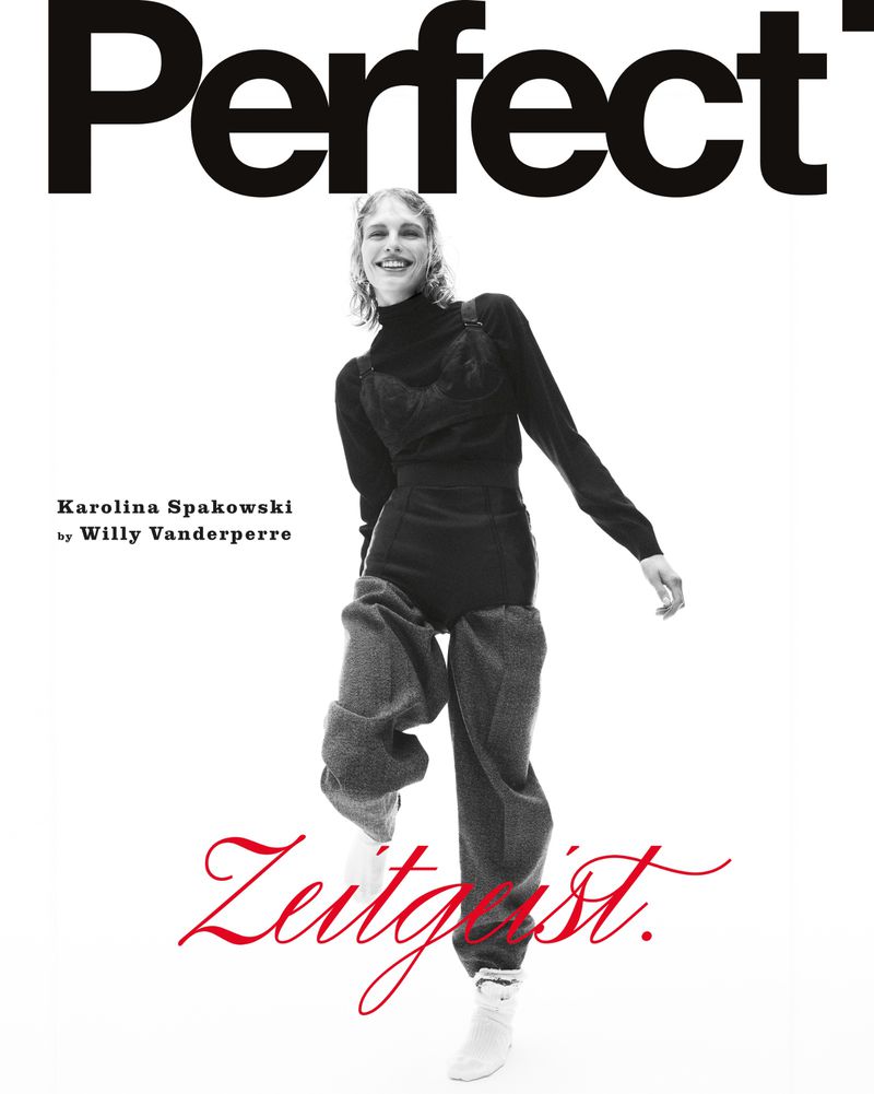 Model Karolina Spakowski posing dynamically in a black top and grey trousers on a white studio background.