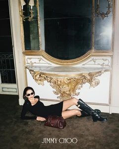 Model wearing a black dress and boots lying on the floor under an ornate gilded console table for Jimmy Choo Fall 2026 Campaign