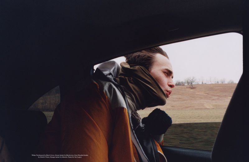 Man in an orange winter jacket and olive neck gaiter leaning his head out of a dark car window toward an open field.