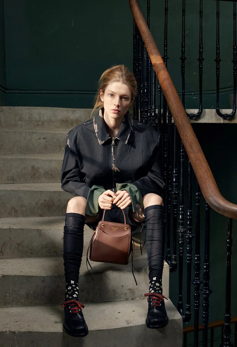 Hunter Schafer sitting on concrete stairs holding a brown leather handbag, wearing an oversized black jacket and dark knee socks.