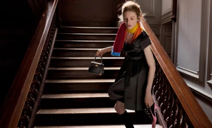 Model in a black dress descending wooden indoor stairs, wearing a colourful striped scarf and carrying a small handbag.