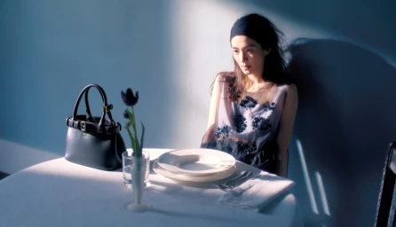 Woman seated at a white-clothed table with a handbag and vase of flowers in a dramatic cinematic light setting for Prada.