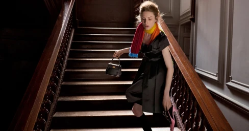 Woman walking down a wooden staircase wearing a dark dress, multi-colored scarf, holding a black handbag.
