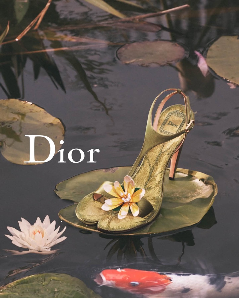 Green stiletto sandal resting on a lily pad in a pond with water lilies and a koi fish underneath.