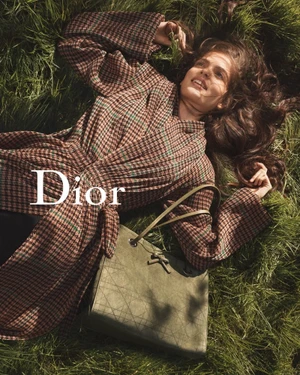 Model lying in tall grass wearing a plaid coat next to an olive green leather handbag for Dior campaign.