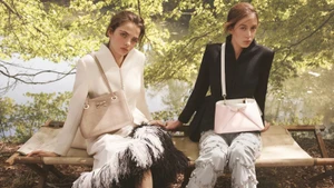 Two models seated outdoors on a rustic wooden bench wearing tailored jackets and holding designer handbags.