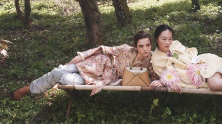 Two female models reclining on a wooden garden bench outdoors in a wooded forest setting wearing patterned coats and holding a structured handbag